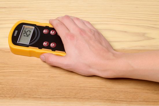 A hand placing a pinless moisture meter on a floorboard to measure its moisture content