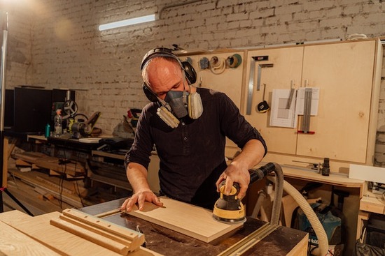 A man wearing a dust mask as he uses an orbital sander on a piece of wood