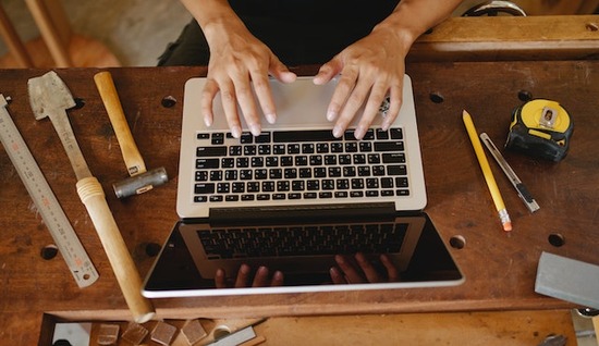 A carpenter using a laptop at his workbench surrounded by tools