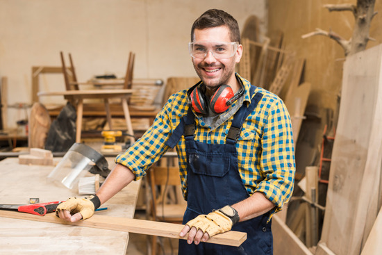 A carpenter holding a wooden plank on a workbench in his shop
