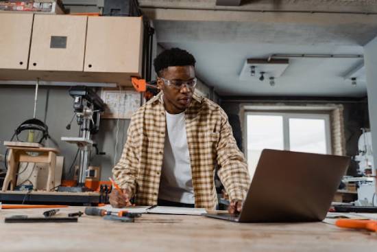 A woodworker in his workshop taking notes as he watches a YouTube video on his laptop
