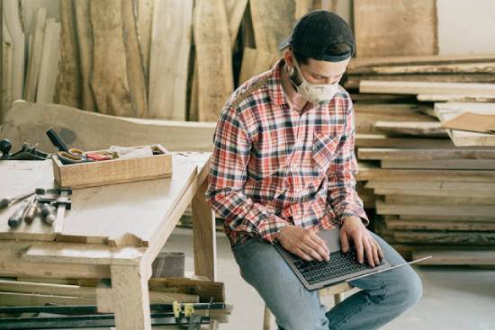 A woodworker in his workshop, wearing a safety mask as he searches for YouTube channels on his laptop