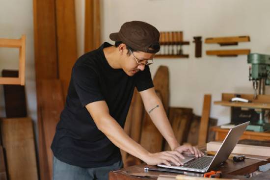 A man standing in his workshop, typing on his laptop as he searches for woodworking YouTube videos