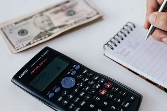 A person making a budget with a notepad and calculator, with cash sitting in the background