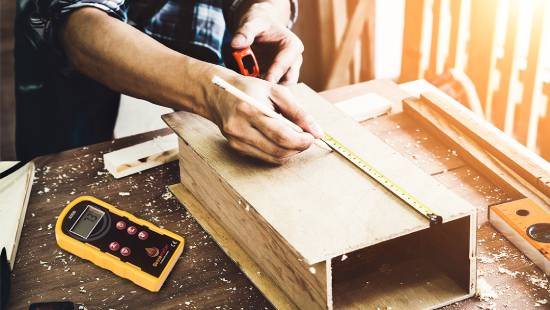 Bessemeter on Table Beside Woodworking Project A man with moisture meter sitting next to him carefully measures a wooden box with a tape measure—demonstrating the importance of accuracy in every aspect of a project.